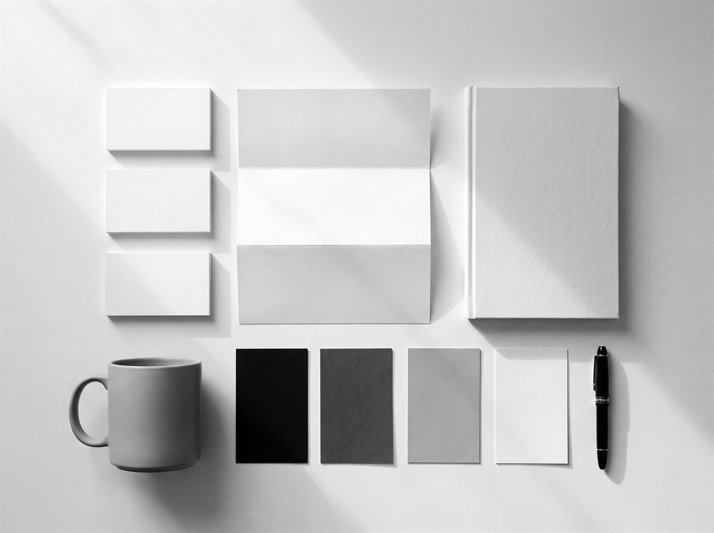 Black-and-white flat lay of a blank brand identity system: business cards, letterhead, notebook, mug and grey swatch cards